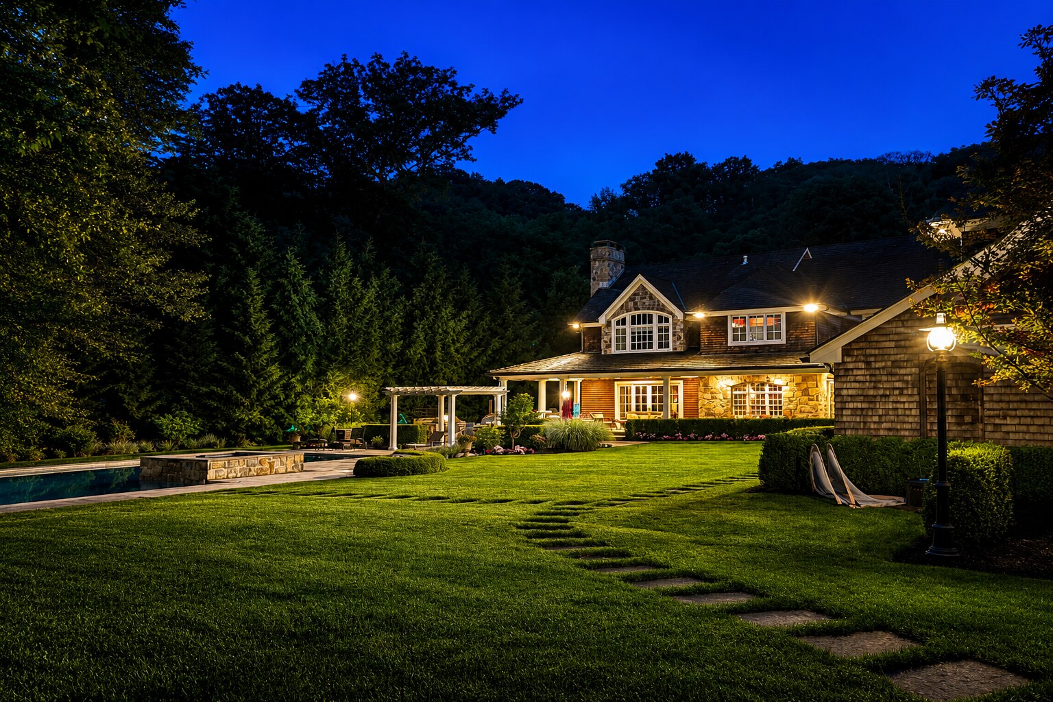 Illuminated stone and shingle estate home with pool, pergola, and a stepping stone path through the lawn at twilight
